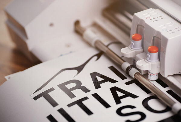 White plotting machine on wooden surface cuts adhesive lettering from black vinyl foil in bright light.